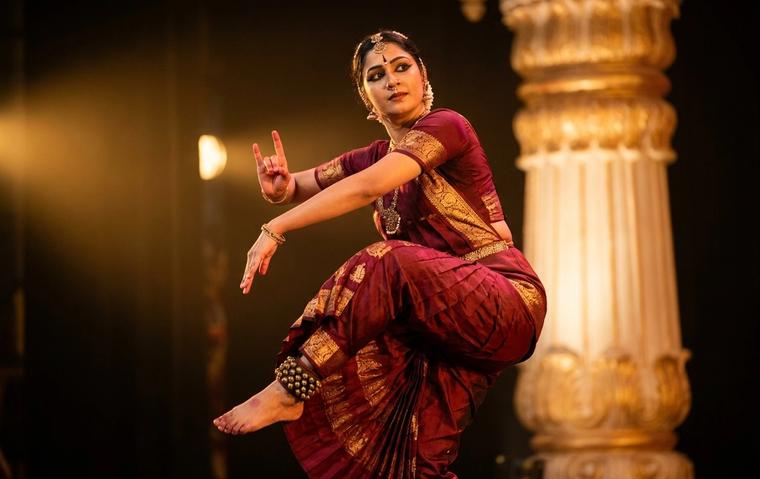 Bharatanatyam dancer in maroon and gold costume holding a sculpturesque thillana pose on a warmly lit stage