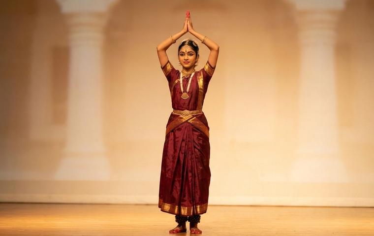 Bharatanatyam dancer in maroon and gold costume standing with palms joined above the head in anjali hasta at the start of an alarippu