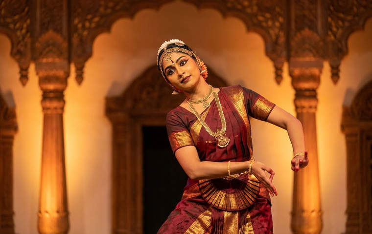A Bharatanatyam dancer in maroon and gold demonstrating a graceful neck movement of griva bheda on a warm, softly lit stage.
