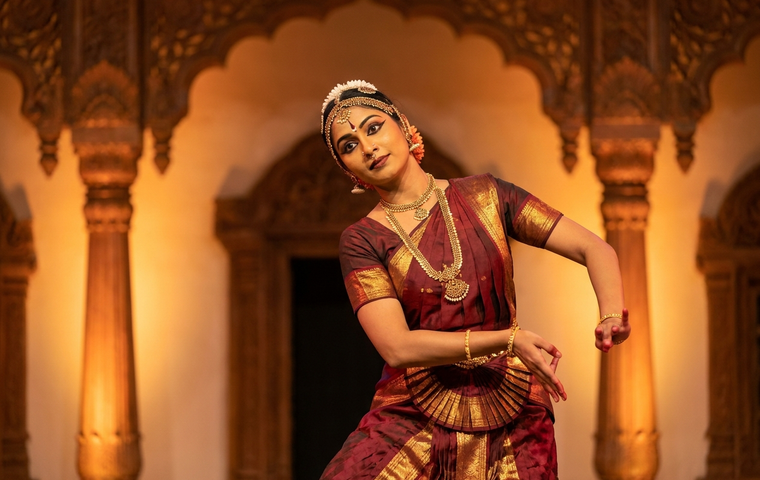 A Bharatanatyam dancer in maroon and gold demonstrating a graceful neck movement of griva bheda on a warm, softly lit stage.