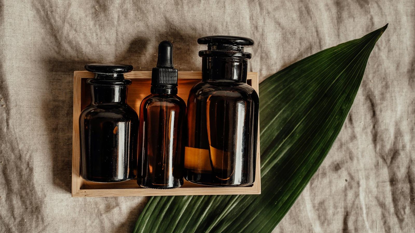 Tres frascos de cosmética de ámbar oscuro dispuestos en una bandeja de madera sobre lino con una hoja de palma