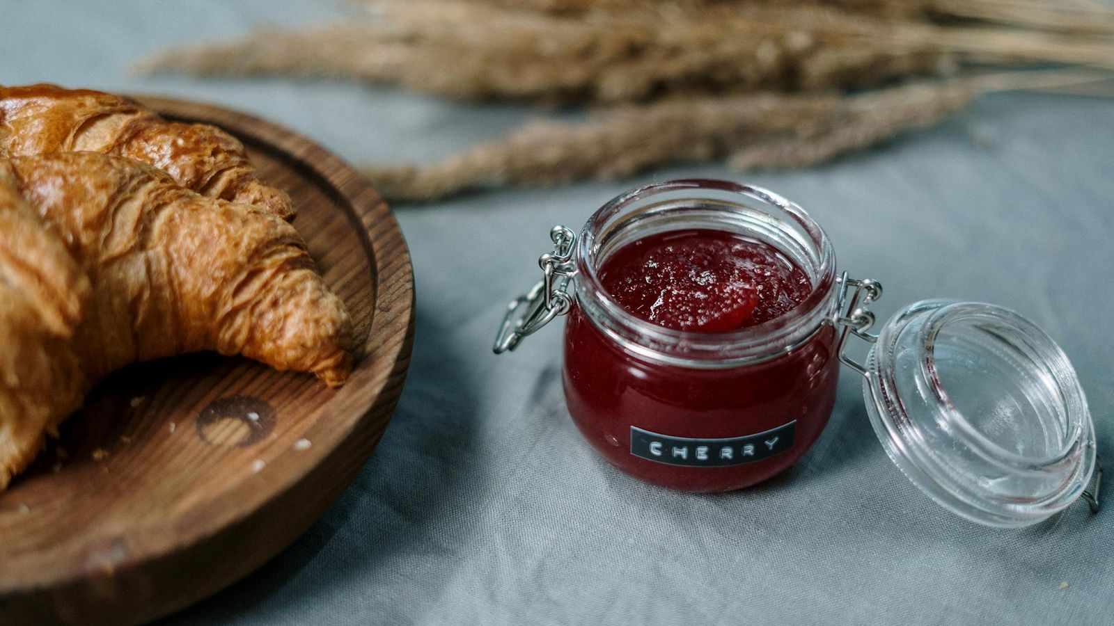 Compota de cereja num pequeno frasco de vidro ao lado de um croissant fresco numa tábua de madeira