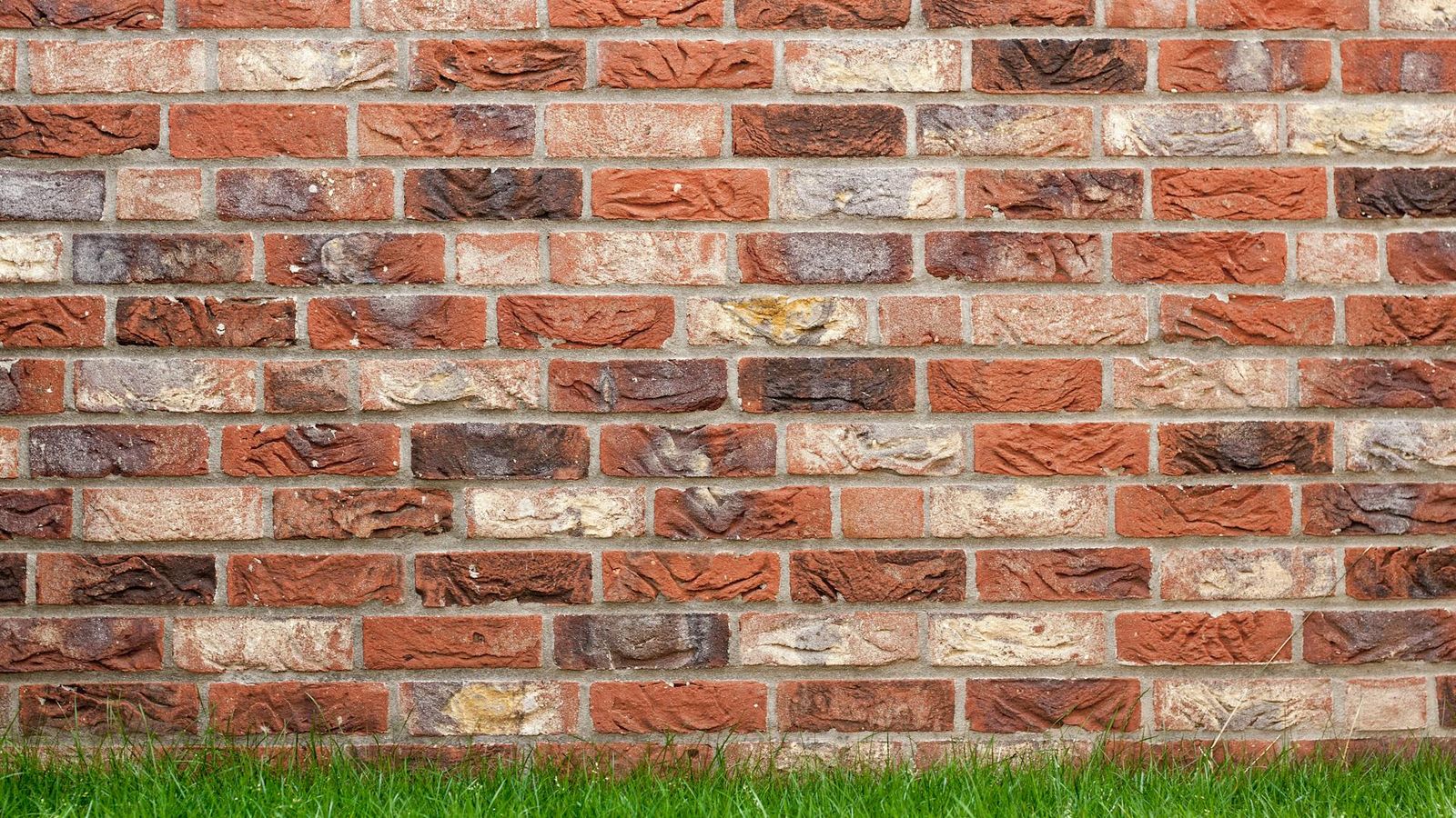 Primo piano di un muro di mattoni rossi e color sabbia con tonalità chiazzate ed erba alla base