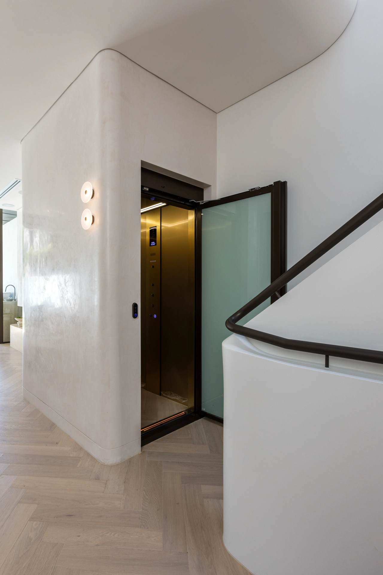 Open brass coloured home lift beside a curved stair wall and dark handrail