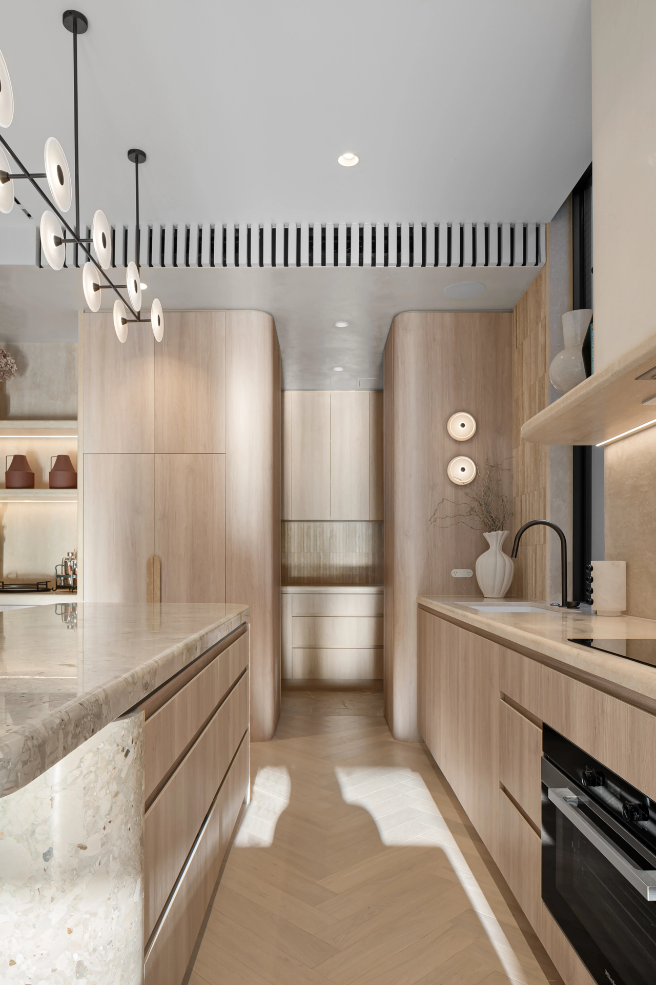 Kitchen with curved timber cabinetry, veined stone benchtops and sunlight across herringbone flooring