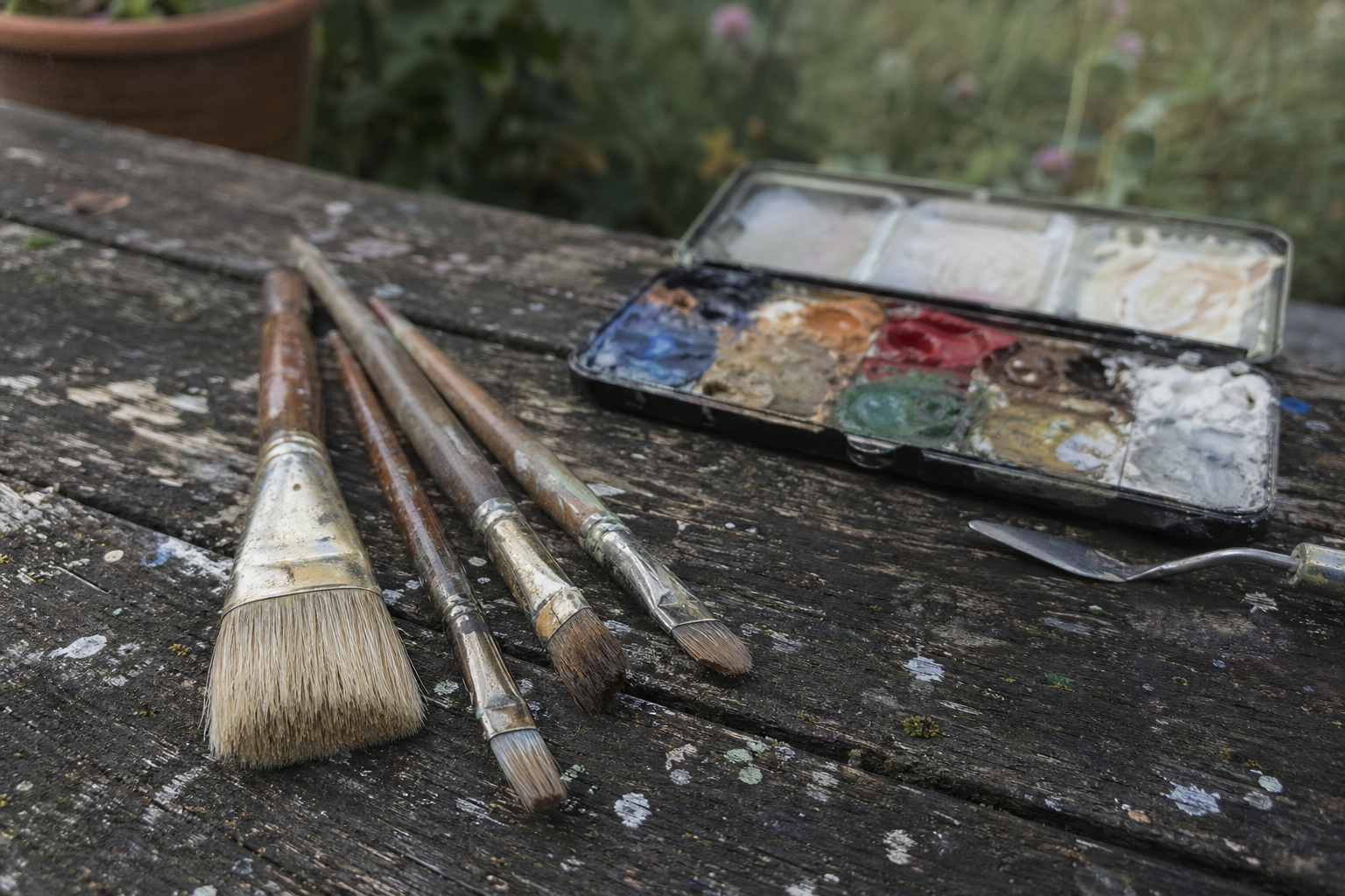 Four oil painting brushes beside a compact palette on a wooden outdoor surface