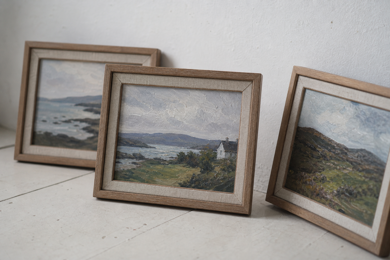 Three small framed landscape paintings leaning against a white wall in natural light