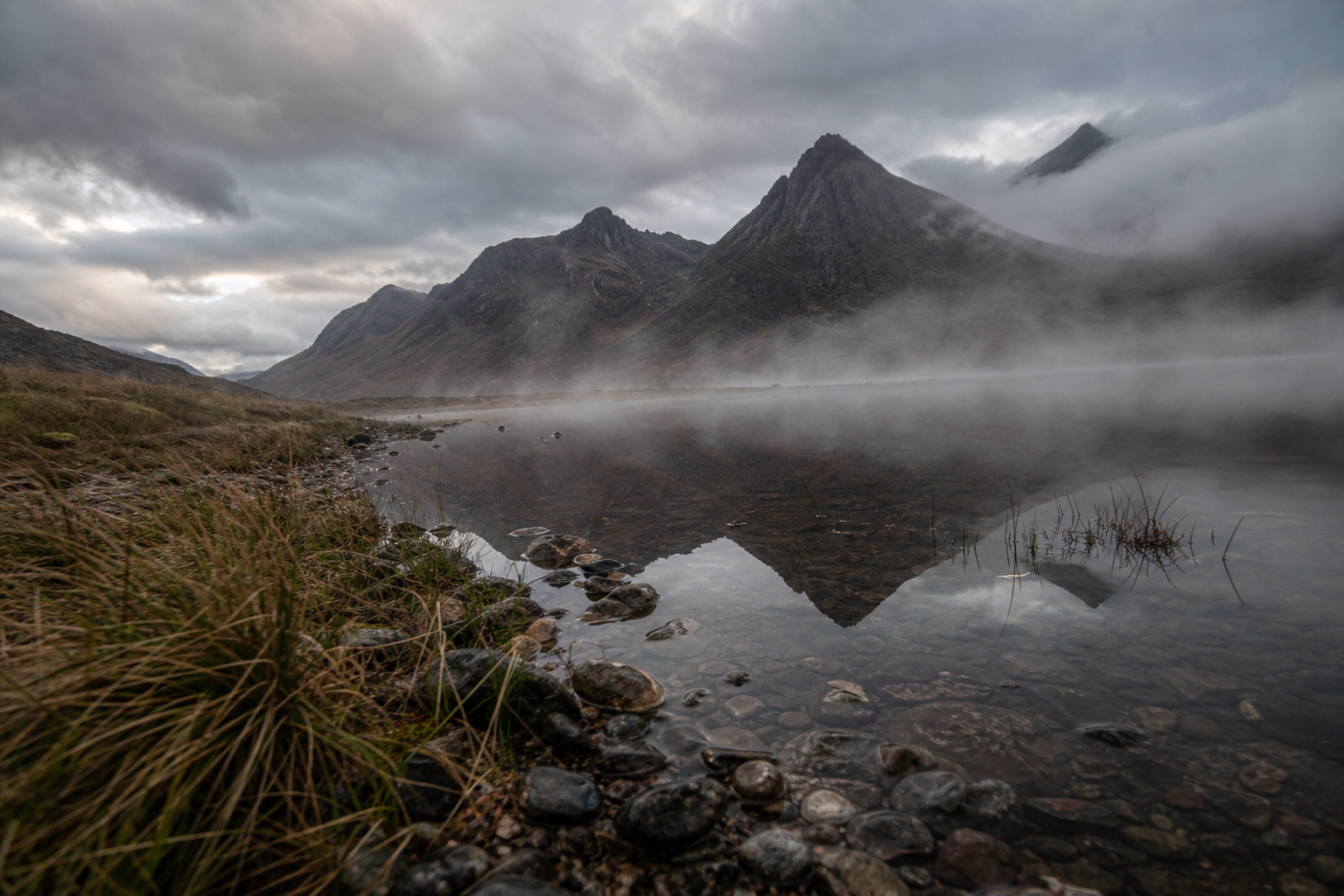 Misty Highland loch reflecting jagged mountain peaks under a dramatic cloudy sky