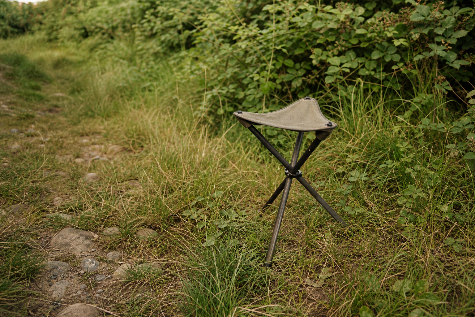 Compact tripod stool on rough grassy ground near a hedgerow