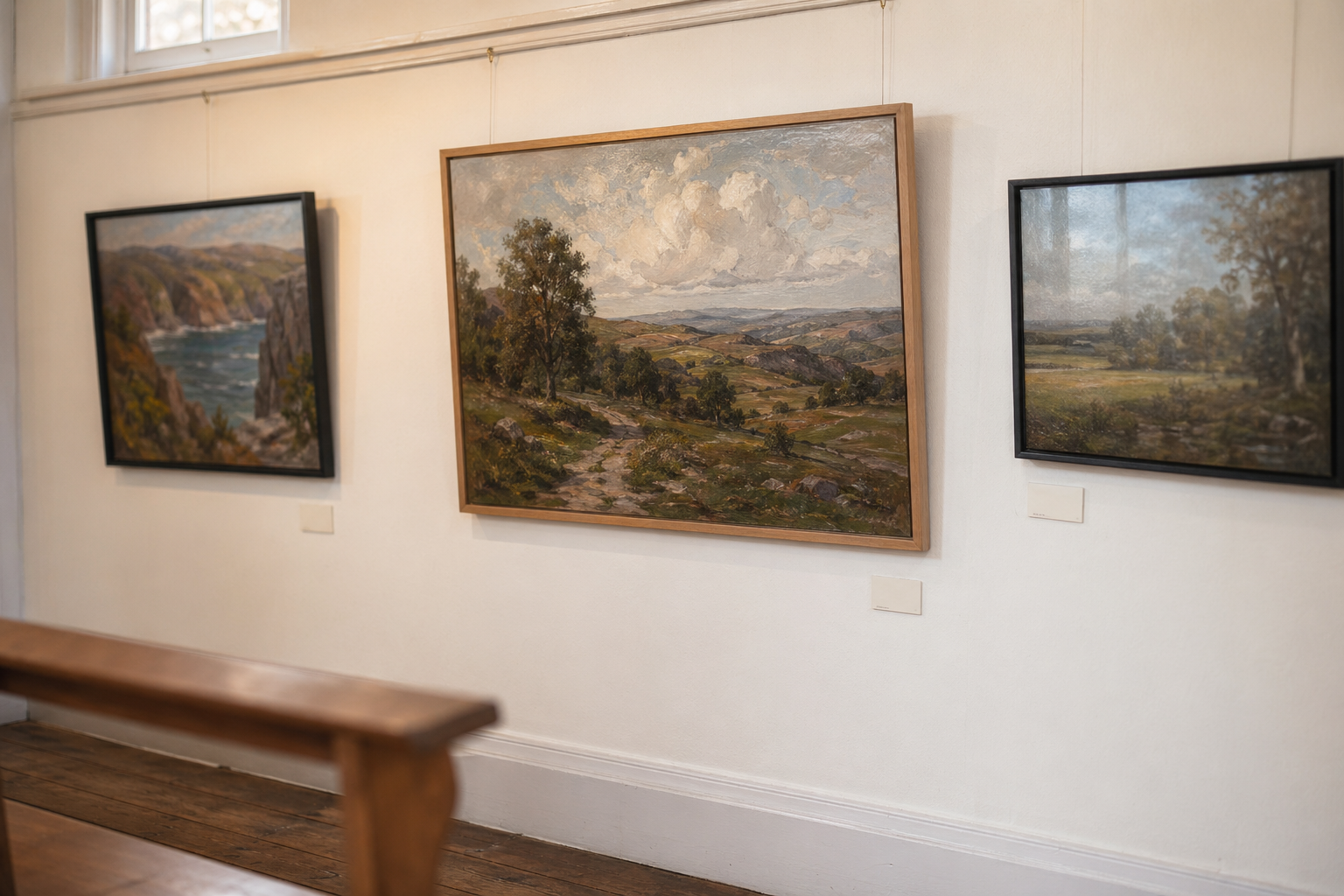 Framed landscape paintings hung on a white gallery wall with warm lighting