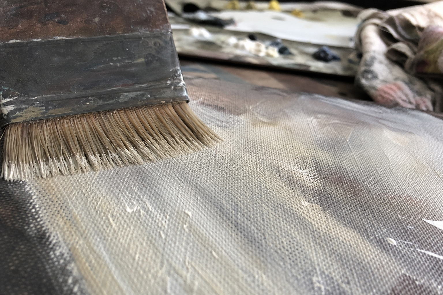 Stiff hog bristle brush dragging pale paint across textured canvas surface