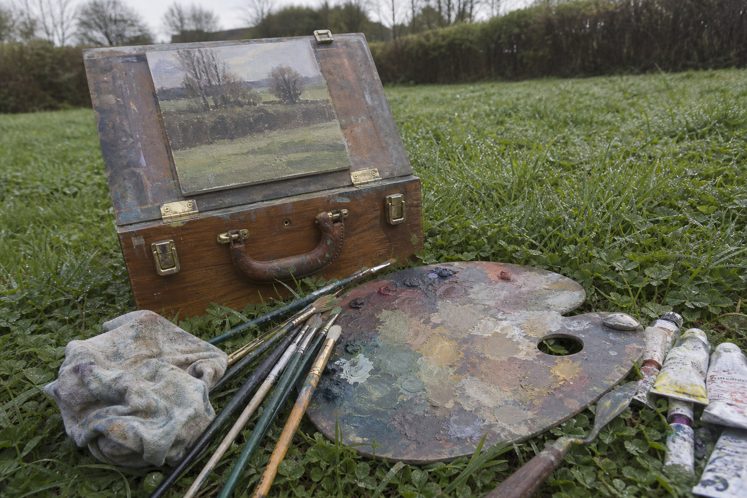 Palette, brushes, and paint tubes arranged on grass beside a pochade box