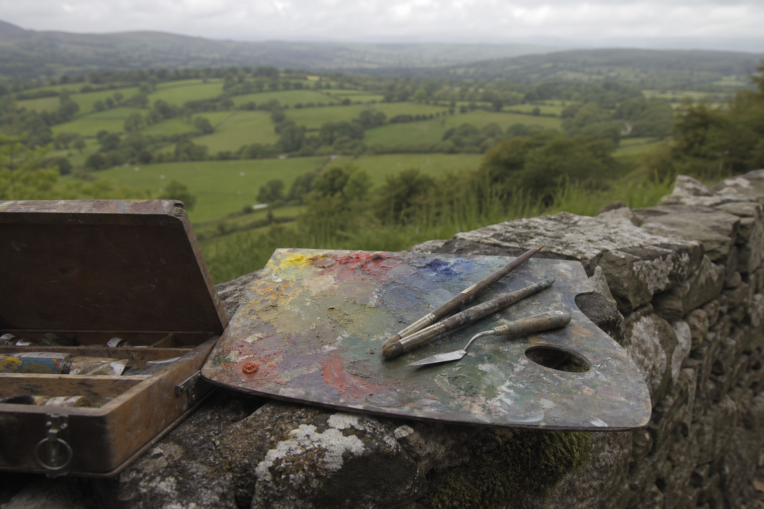 Paint box and palette resting on a dry stone wall overlooking a green Devon valley