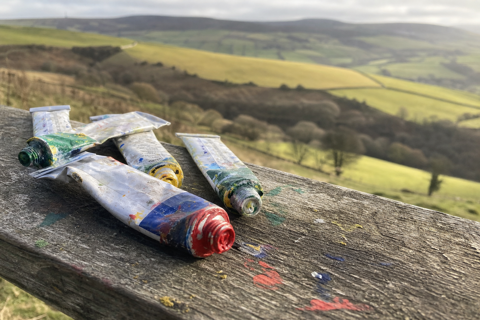 Colourful oil paint tubes arranged on rough wood, rolling British hills visible beyond