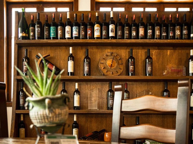 Rows of wine bottles on a tiered wooden display shelf in a rustic dining room, with a potted plant and wooden chairs in the foreground.