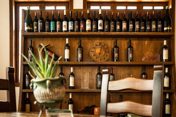 Rows of wine bottles on a tiered wooden display shelf in a rustic dining room, with a potted plant and wooden chairs in the foreground.