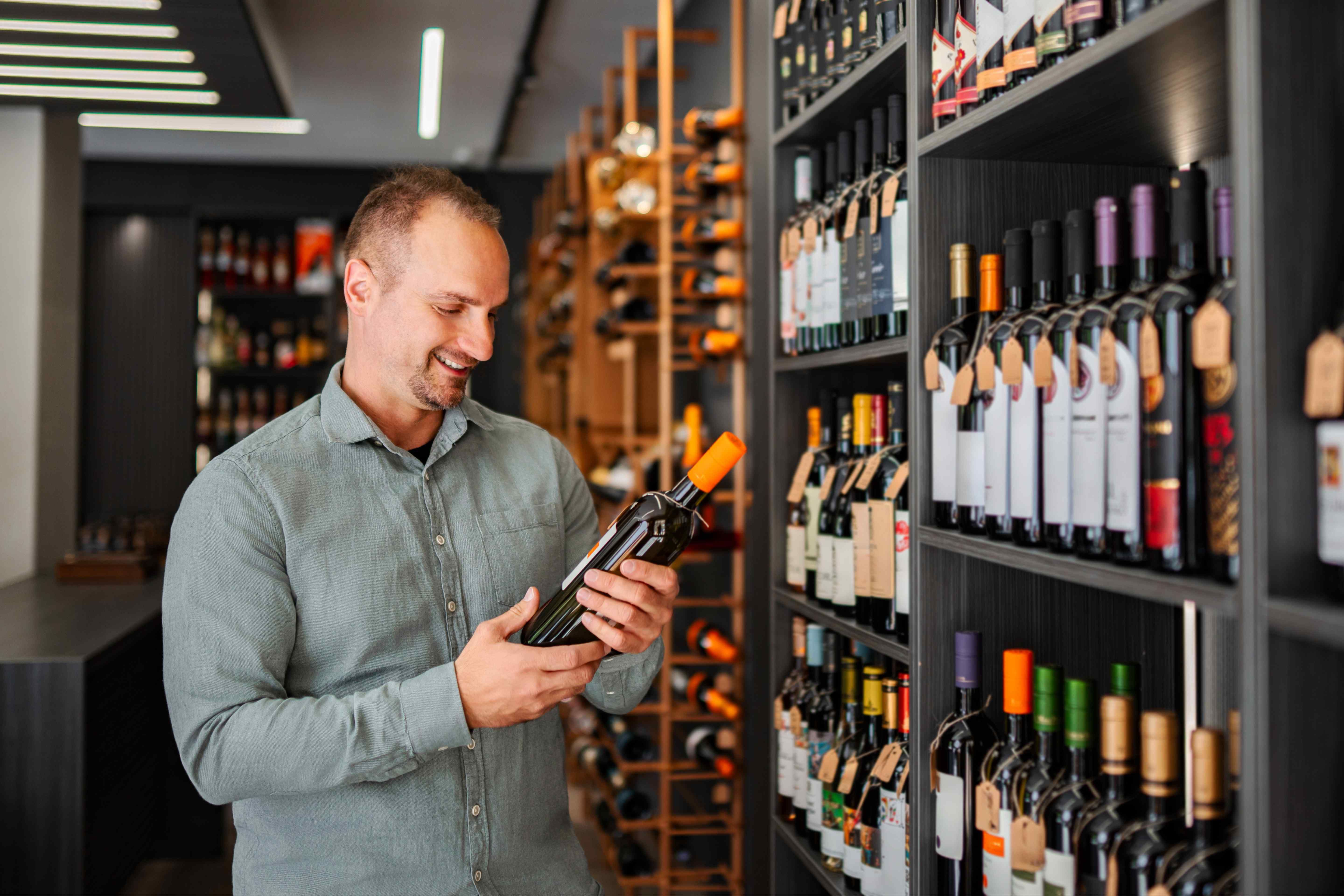 Man examining a wine bottle in a wine shop with fully stocked shelves of tagged bottles behind him.