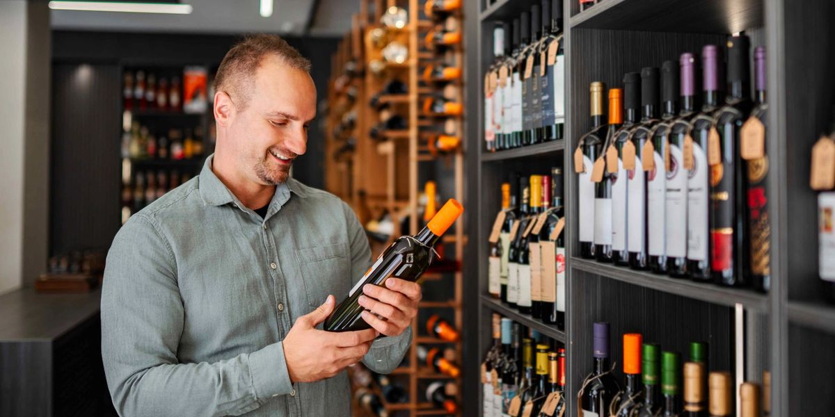 Man examining a wine bottle in a wine shop with fully stocked shelves of tagged bottles behind him.