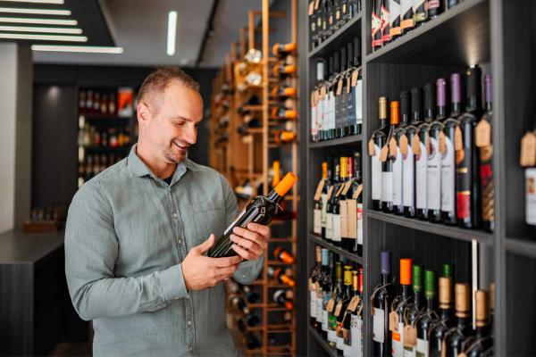 Man examining a wine bottle in a wine shop with fully stocked shelves of tagged bottles behind him.