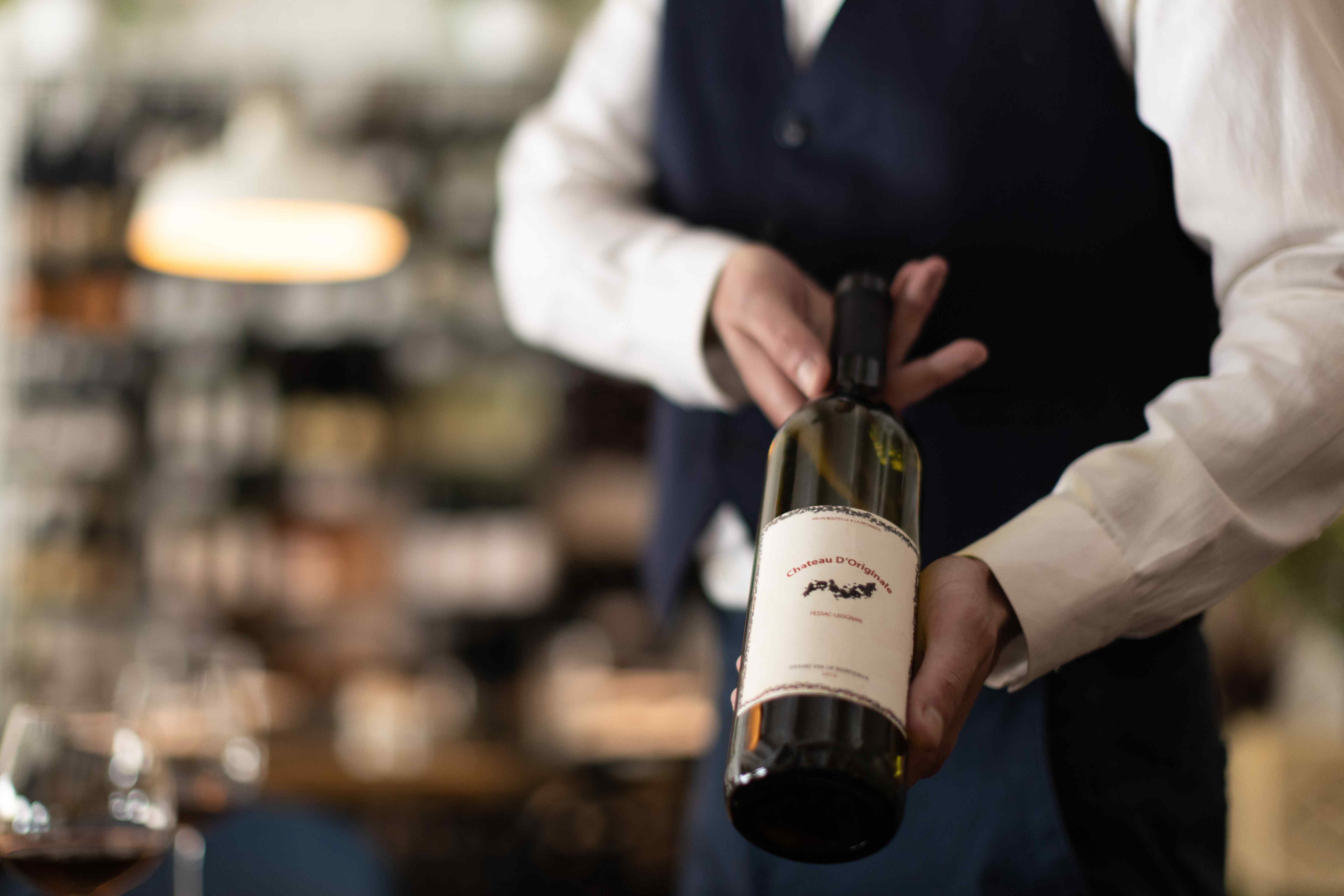 A server in a white shirt and navy vest holds a wine bottle with a cream label in a softly lit restaurant, with a glass of red wine blurred in the foreground.