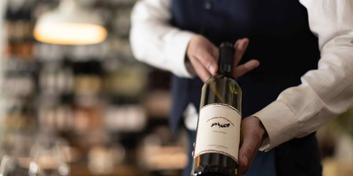 A server in a white shirt and navy vest holds a wine bottle with a cream label in a softly lit restaurant, with a glass of red wine blurred in the foreground.