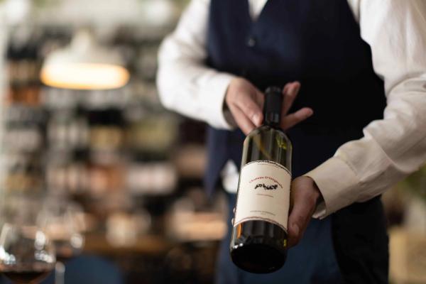 A server in a white shirt and navy vest holds a wine bottle with a cream label in a softly lit restaurant, with a glass of red wine blurred in the foreground.