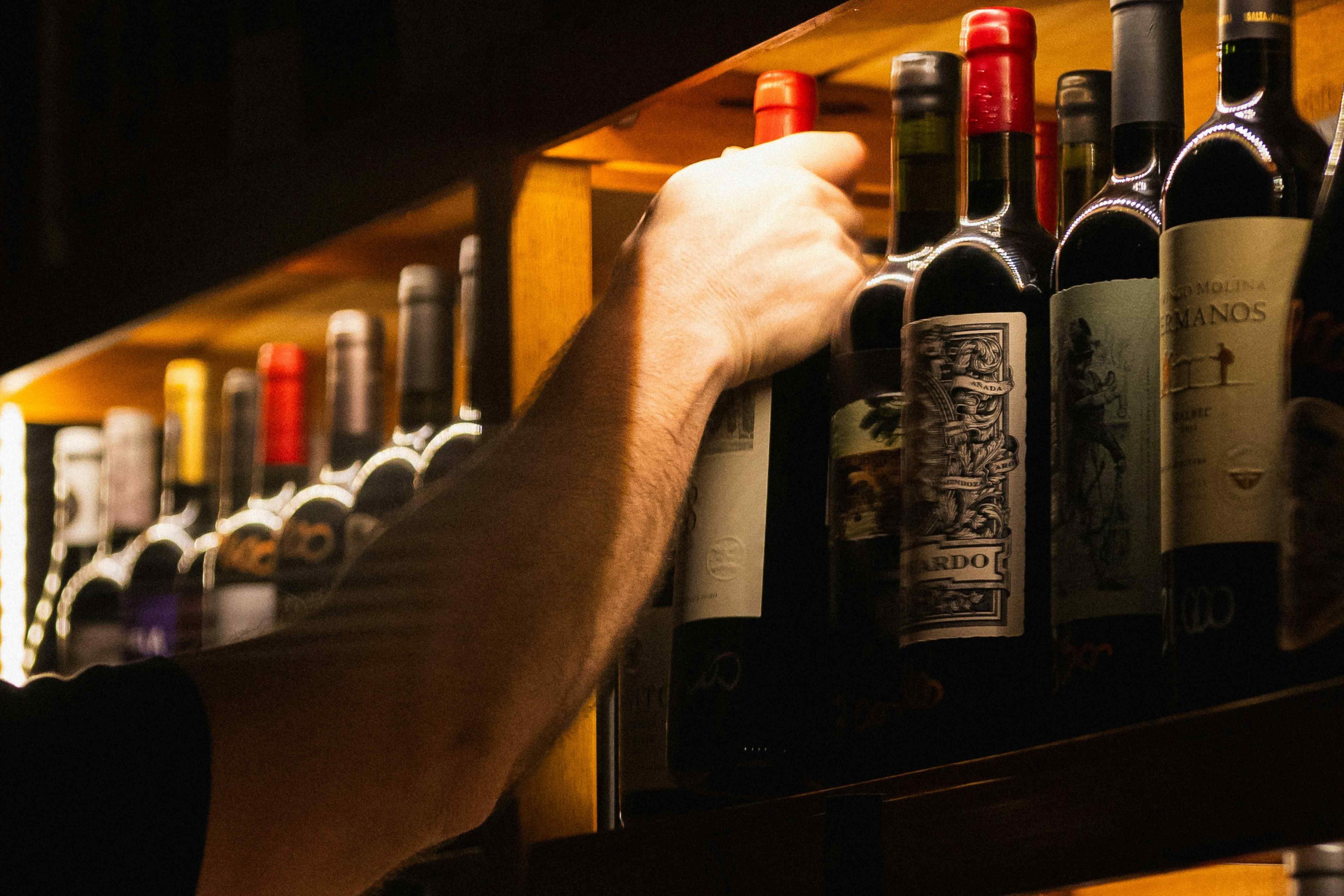 Hand reaching up to pull a bottle from a wooden wine rack lined with red-capped bottles.