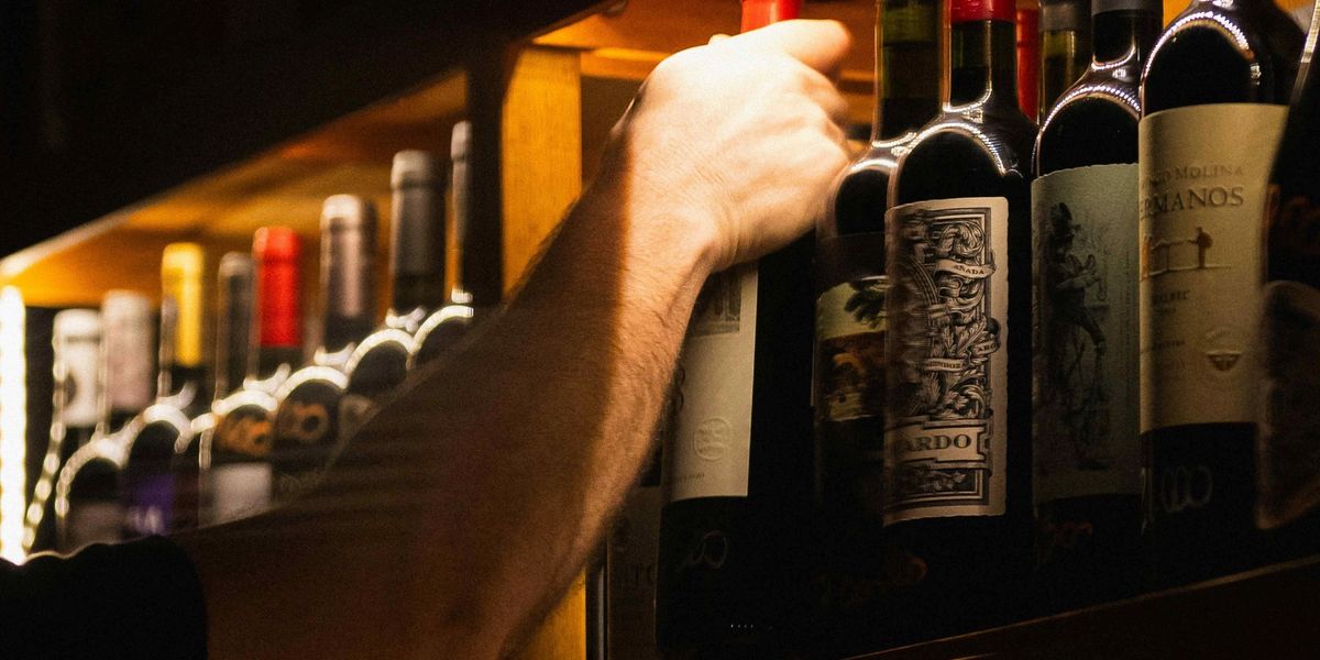 Hand reaching up to pull a bottle from a wooden wine rack lined with red-capped bottles.