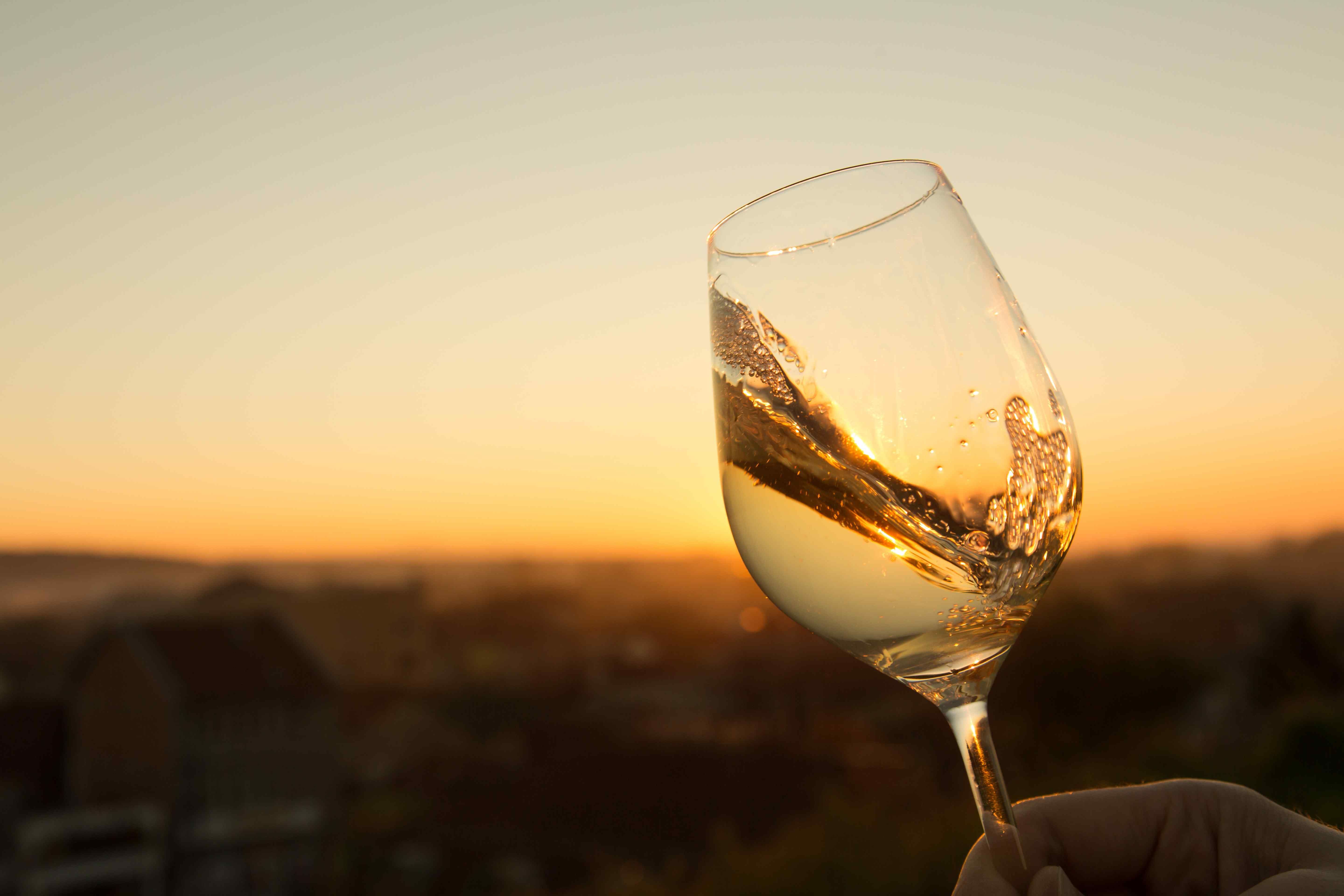A hand holding up a glass of white wine against a golden sunset, with rooftops blurred in the distance below.