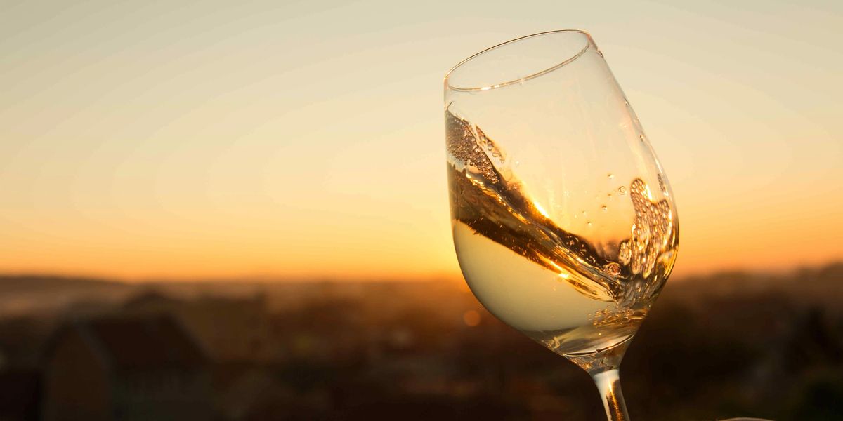 A hand holding up a glass of white wine against a golden sunset, with rooftops blurred in the distance below.