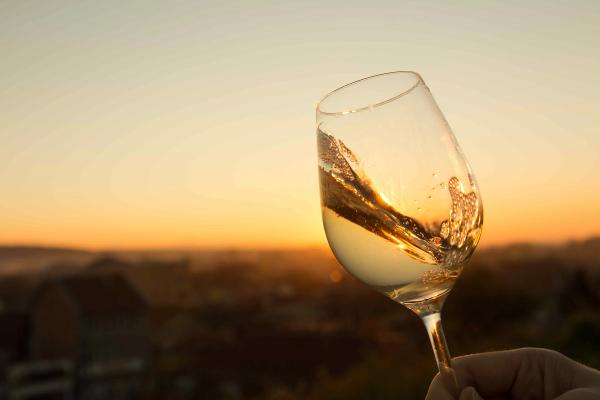 A hand holding up a glass of white wine against a golden sunset, with rooftops blurred in the distance below.