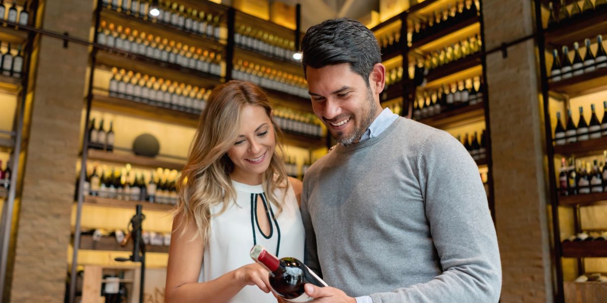A man and woman in a wine shop, looking at a wine label and smiling.