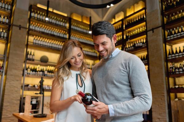 A man and woman in a wine shop, looking at a wine label and smiling.