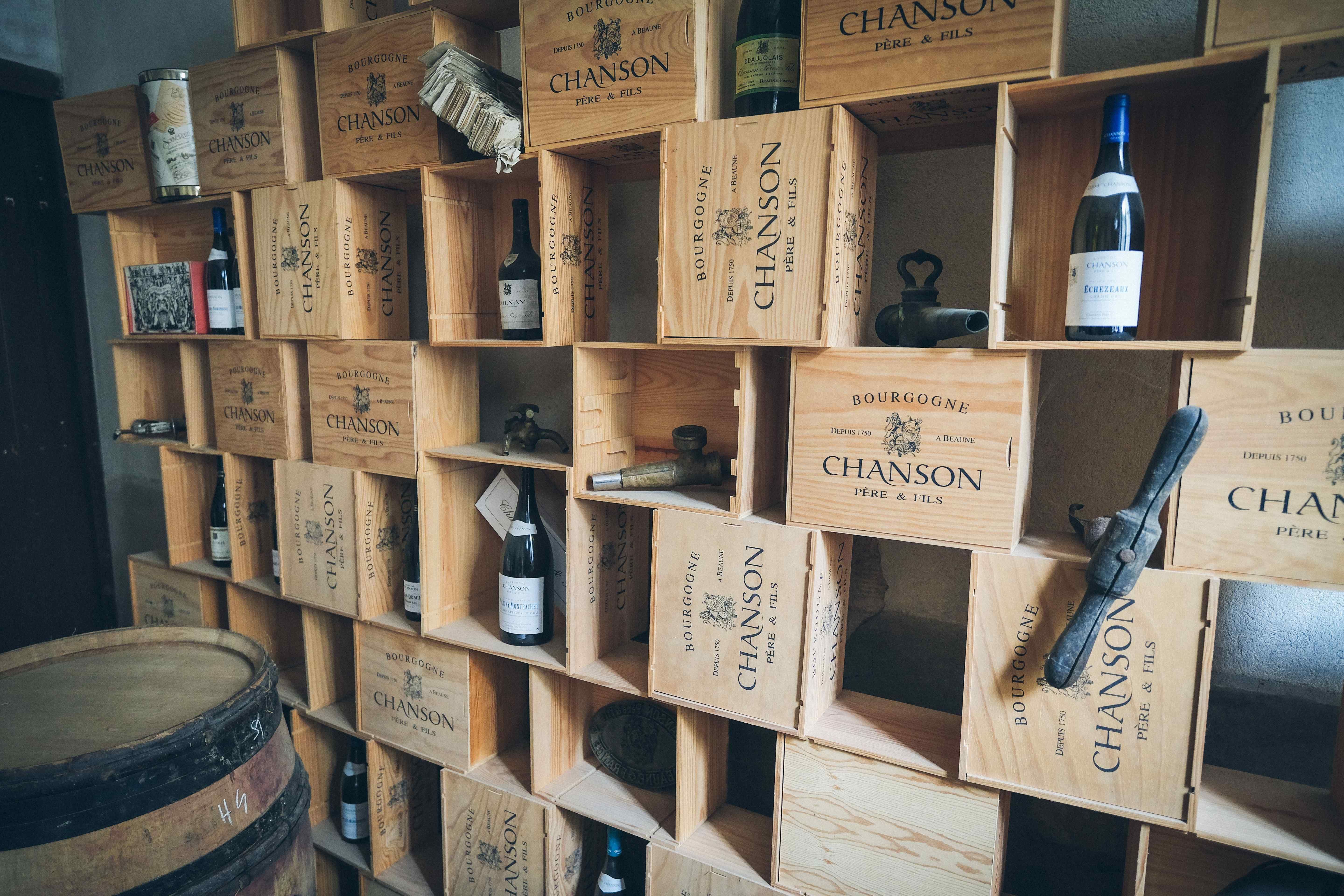 A wall of stacked wooden Burgundy wine cases used as shelving, holding bottles, antique cellar taps and an oak barrel.