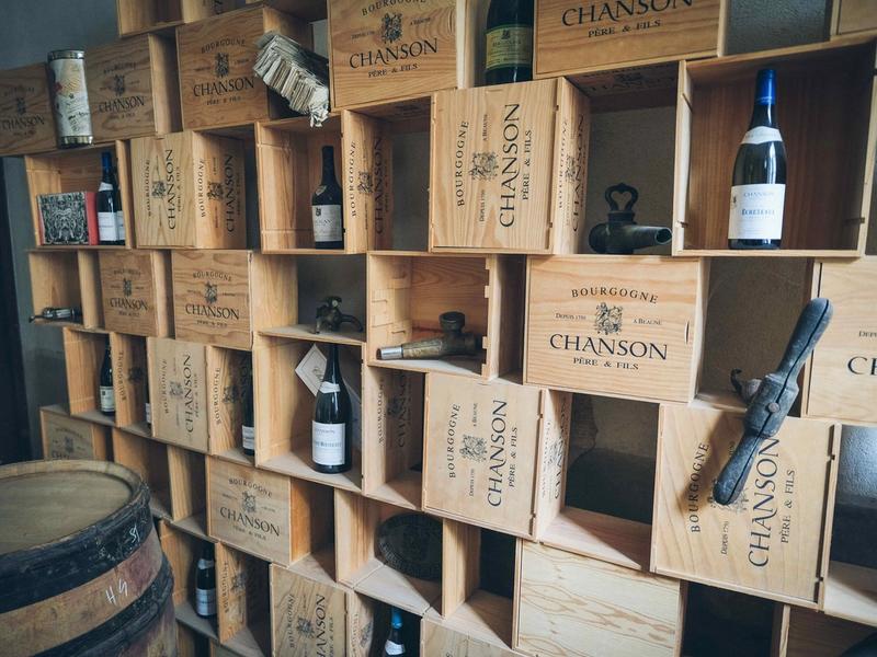 A wall of stacked wooden Burgundy wine cases used as shelving, holding bottles, antique cellar taps and an oak barrel.