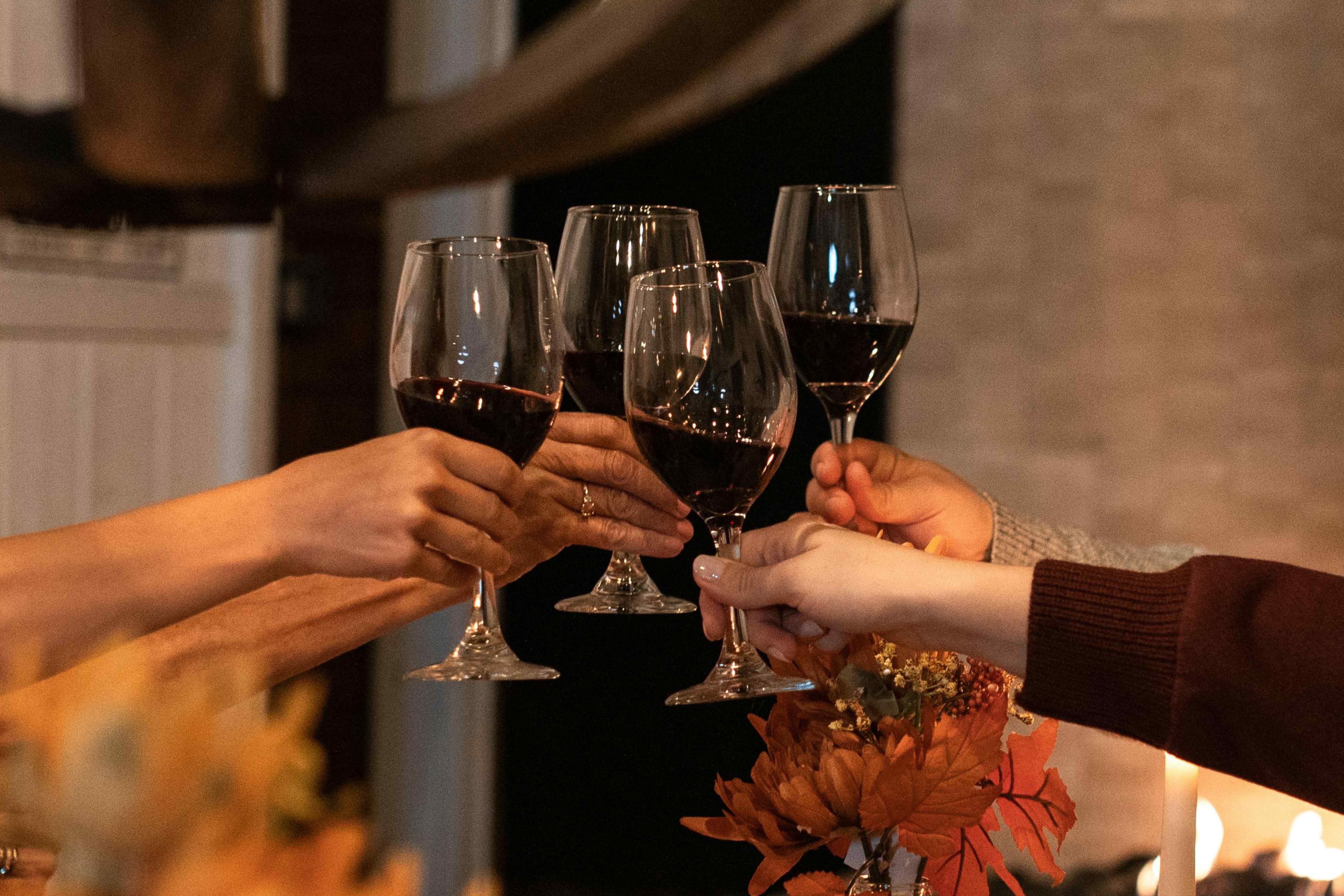 Four hands clinking glasses of red wine together near an autumn floral arrangement.