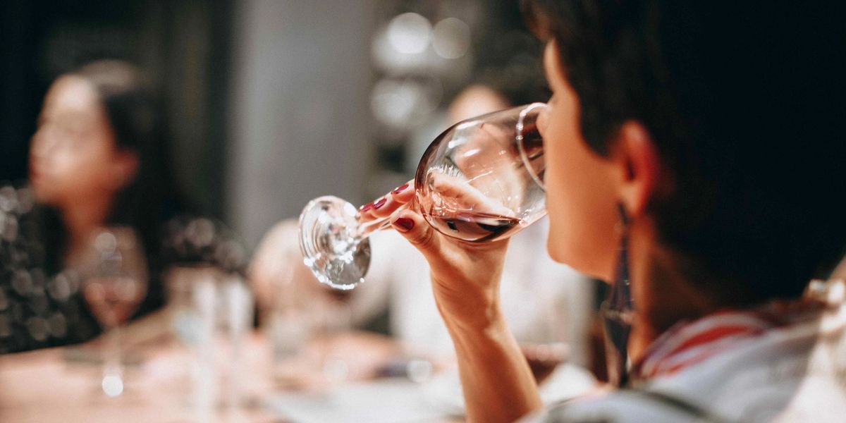 A woman tasting red wine at a dimly lit dinner table with other guests in the background.