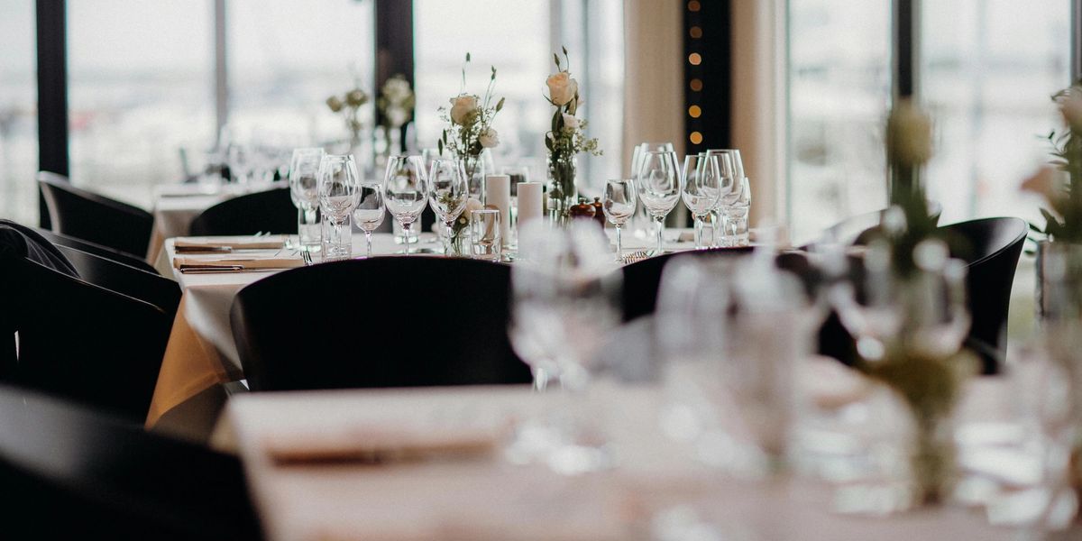 A brightly-lit room full of restaurant tables and chairs, with wine glasses and flowers standing on each table.