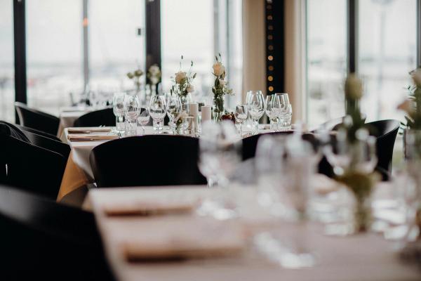 A brightly-lit room full of restaurant tables and chairs, with wine glasses and flowers standing on each table.