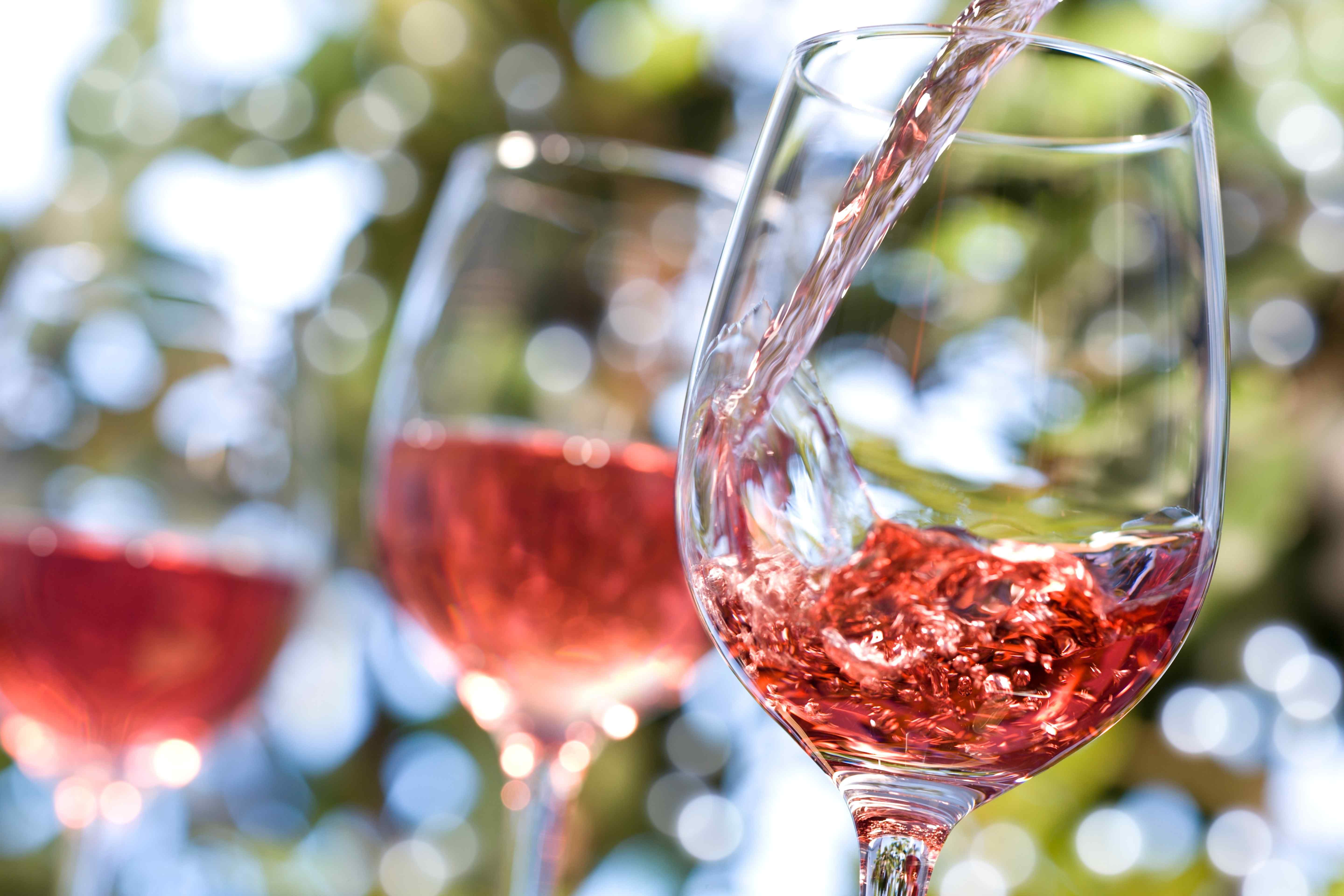 Pale red wine being poured into a stemmed glass outdoors, with two more glasses of the same wine blurred in the background.