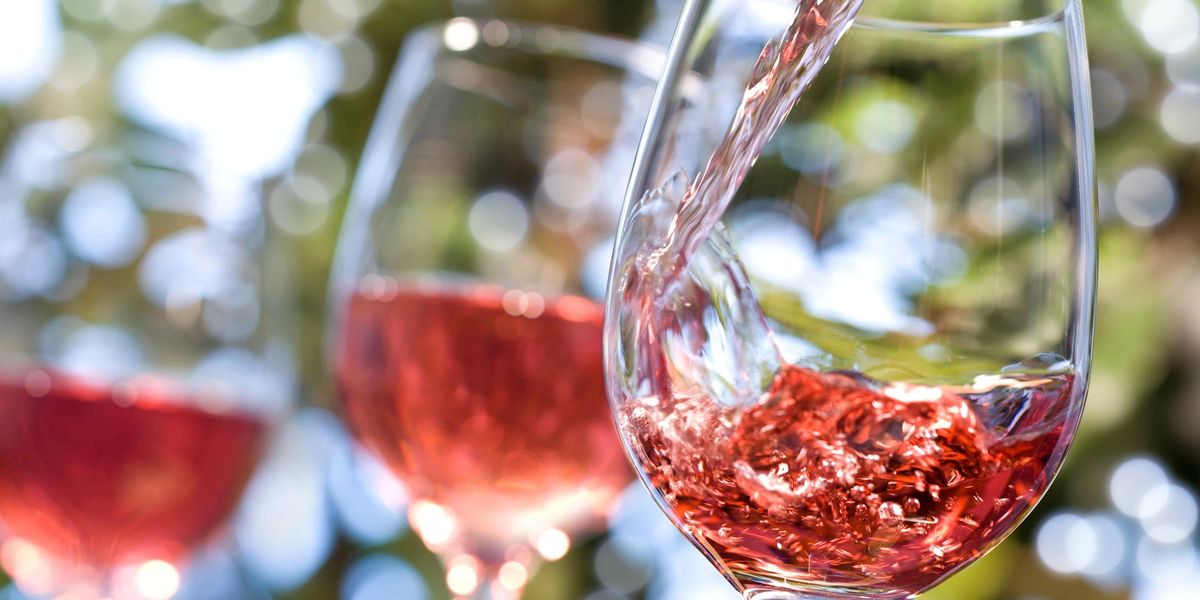 Pale red wine being poured into a stemmed glass outdoors, with two more glasses of the same wine blurred in the background.