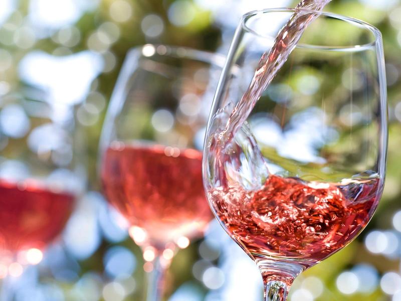 Pale red wine being poured into a stemmed glass outdoors, with two more glasses of the same wine blurred in the background.