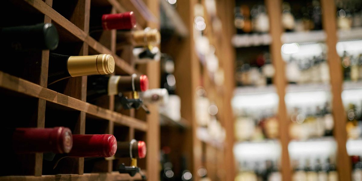 Wine bottles resting in a wooden diamond rack with a blurred cellar of shelved bottles in the background.