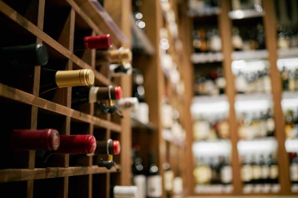 Wine bottles resting in a wooden diamond rack with a blurred cellar of shelved bottles in the background.