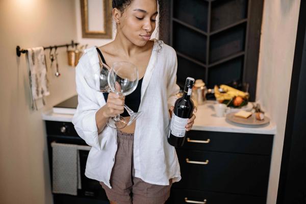 A young woman at home, holding a wine bottle and two empty wine glasses.