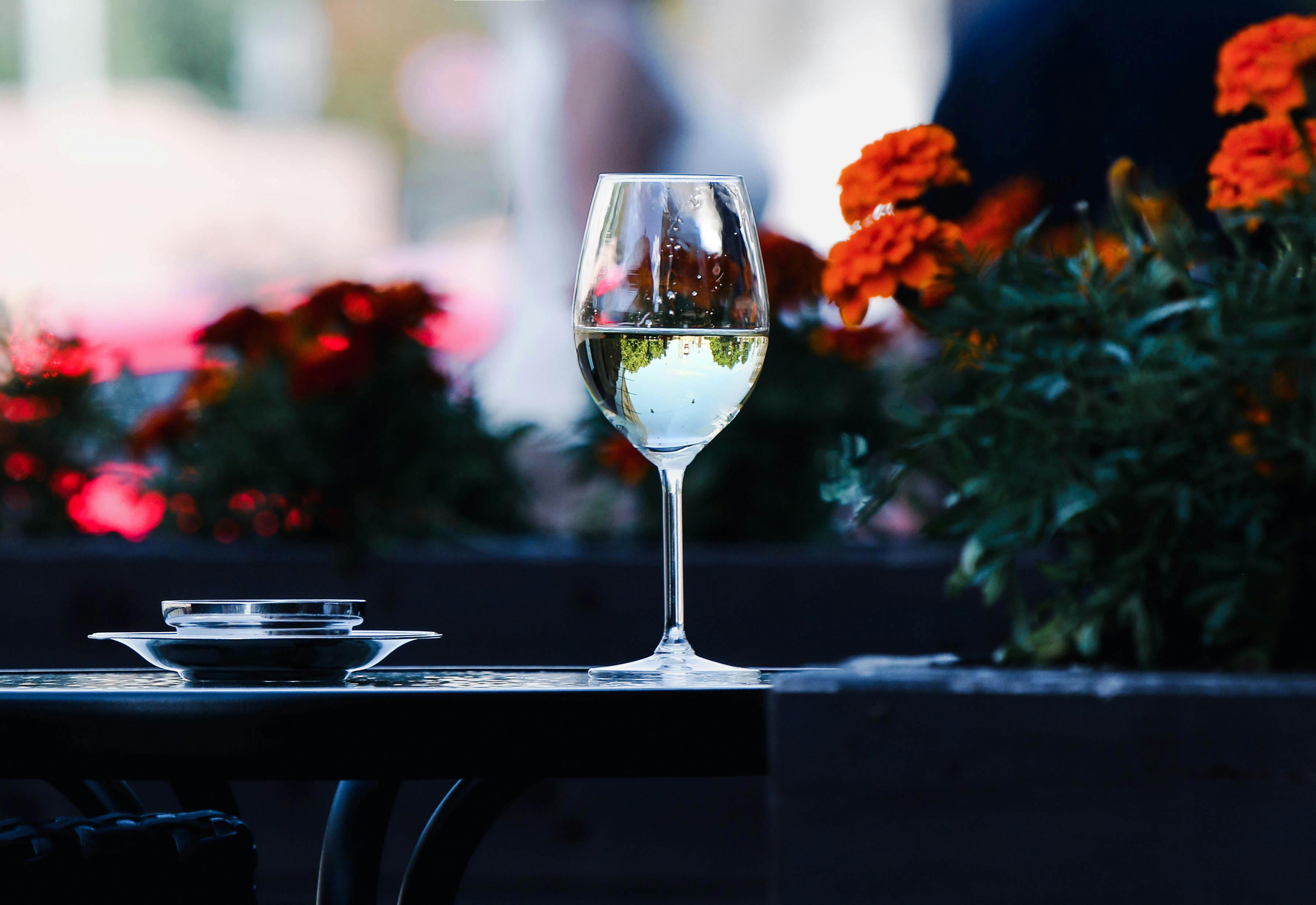 A full glass of white wine rests on a table, with blooming flowers in the background.