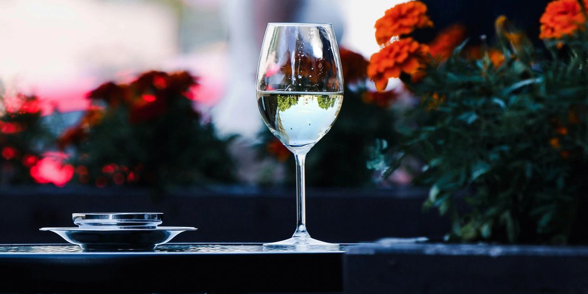 A full glass of white wine rests on a table, with blooming flowers in the background.