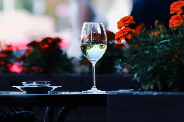 A full glass of white wine rests on a table, with blooming flowers in the background.