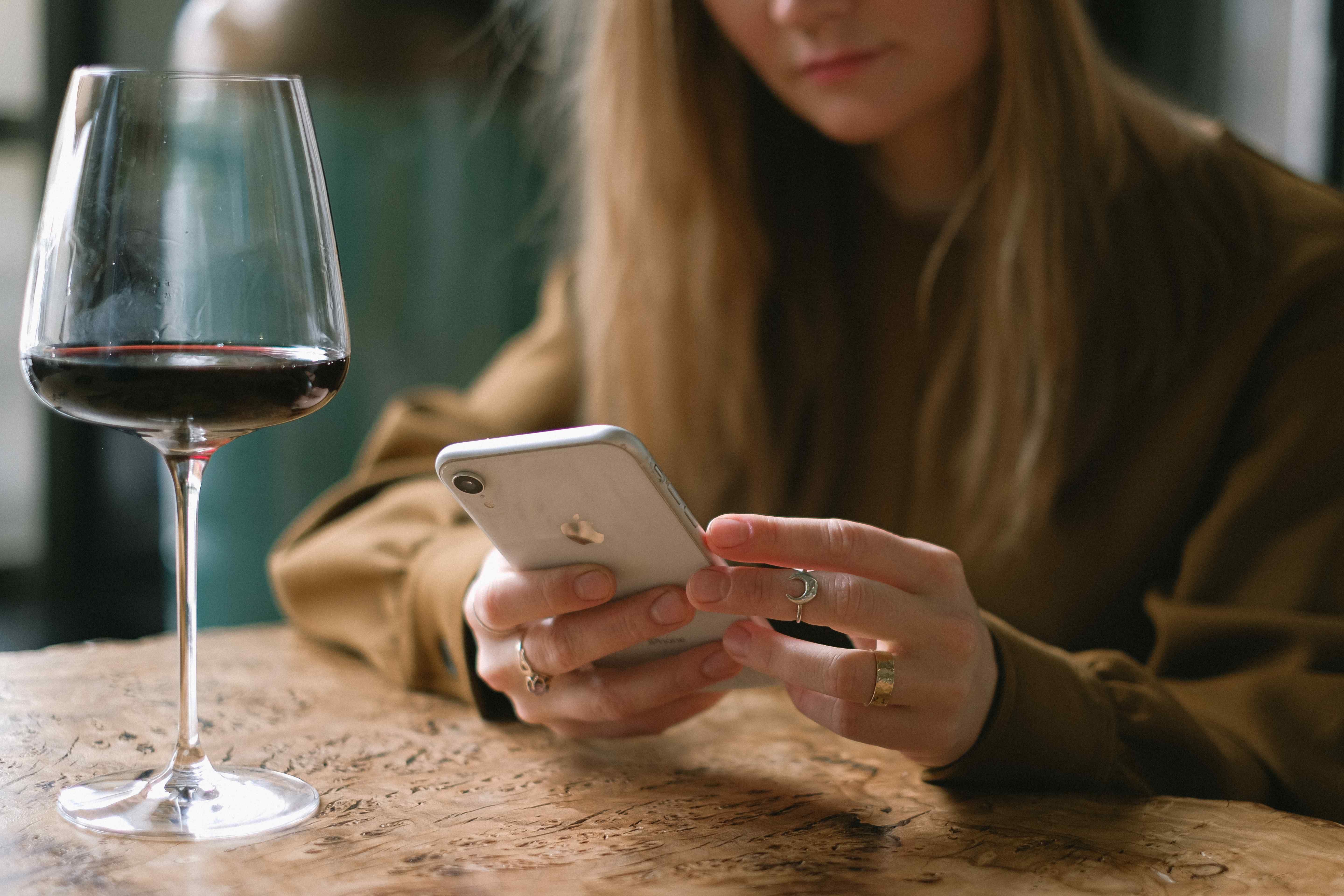 A woman using an iPhone wine app beside a glass of red wine on a wooden table.