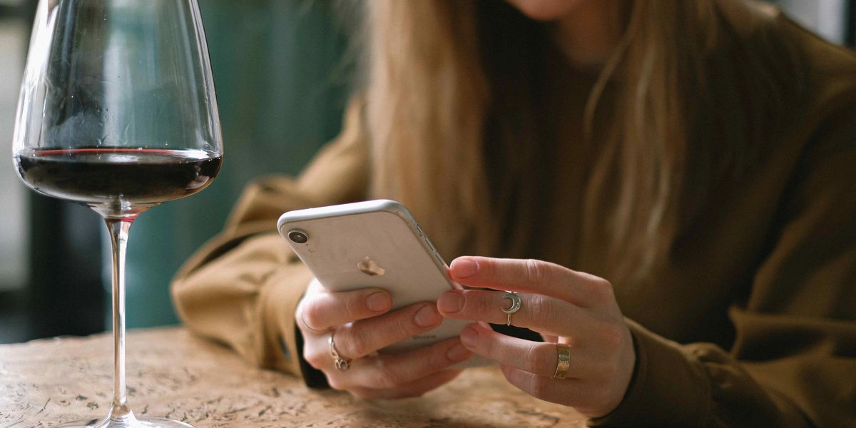 A woman using an iPhone wine app beside a glass of red wine on a wooden table.